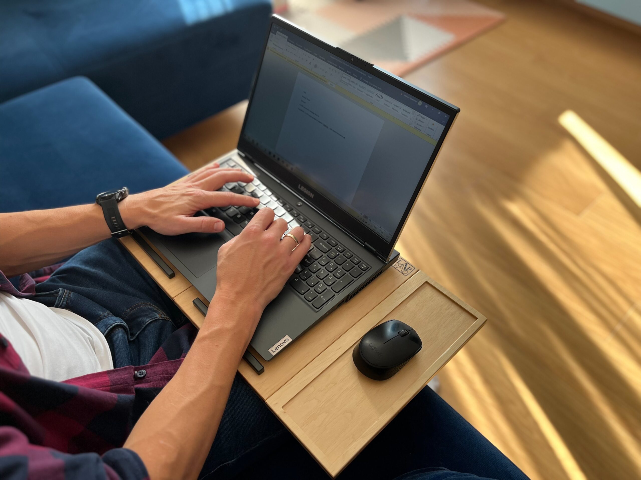 Mężczyzna pracujący na laptopie Lenovo z myszką, korzystający z ergonomicznej podstawki LapPad, siedząc na sofie w jasnym, nowoczesnym wnętrzu.